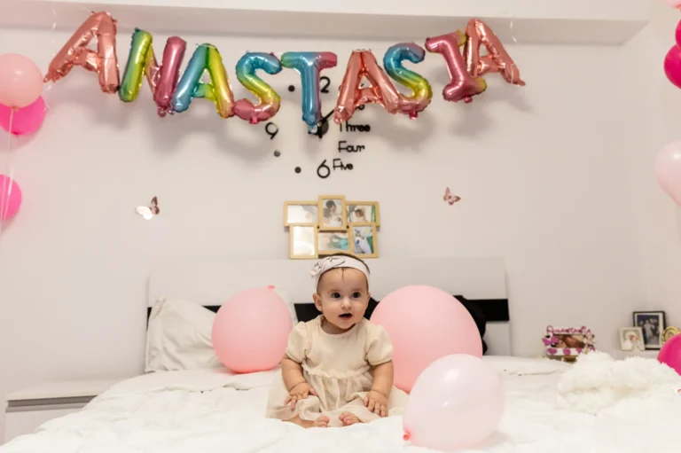 Fetița așezată pe pat, sub baloanele cu numele ei, Anastasia