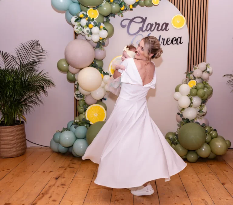 Mama dansează cu fetița în brațe, în fața panoului cu baloane cu numele ei