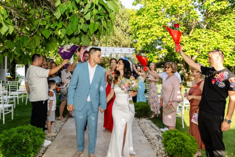 Mirii merg de mână pe aleea din grădină, aplaudați de invitați cu flori