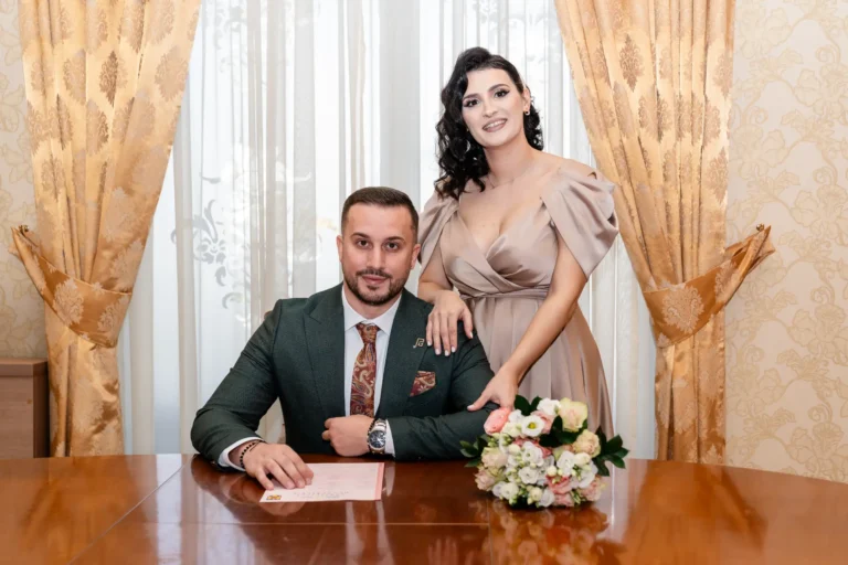 Mirele semnează la masa ofițerului de stare civilă, cu mireasa sprijinită de umărul lui