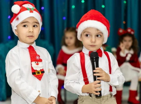 Doi băieței cu căciuli de Moș Crăciun spunând o poezie la microfon