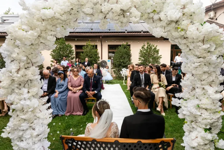 Ceremonie în aer liber, sub o arcadă de flori albe, cu invitații așezați