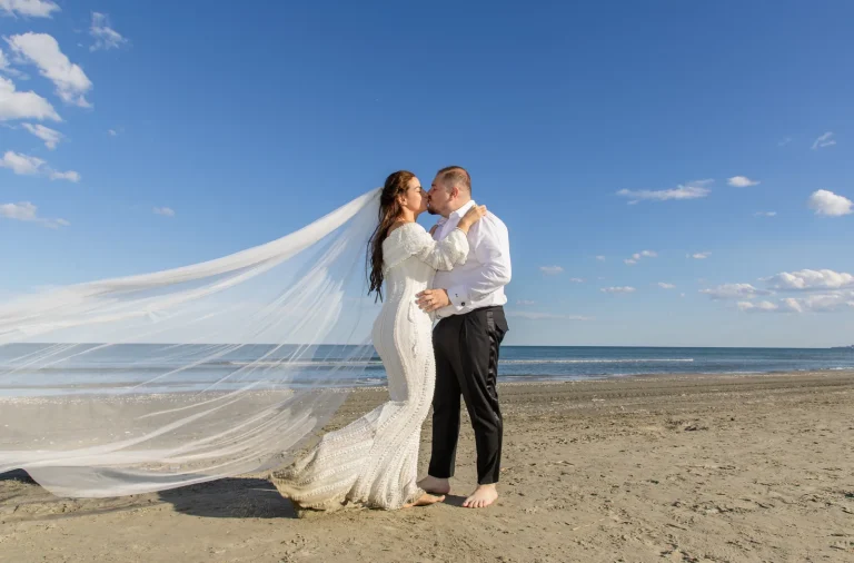 Sărutul mirilor pe plajă, cu voalul miresei purtat de vânt